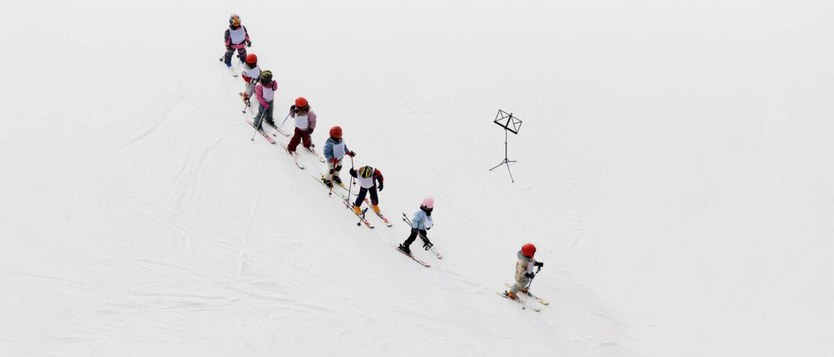 Eine Schlange aus Kindern in Skiausrüstung auf einer schneebedeckten Piste. Daneben ein Notenständer.